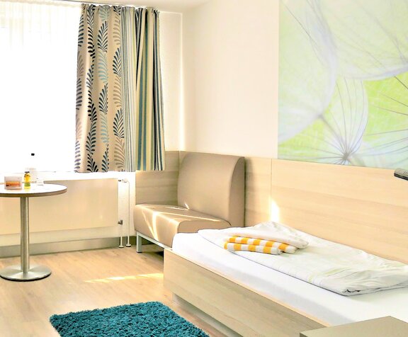 Ausschnitt eines Patientenzimmers mit Bett und einem kleinen Tisch mit Sitzsofa.