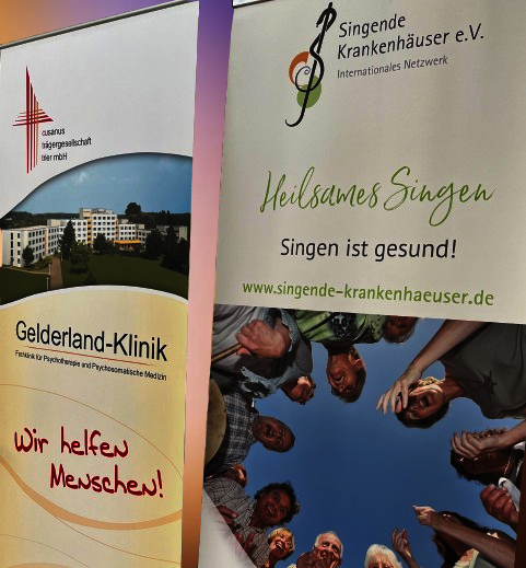 Auf dem Bild sind zwei Aufsteller abgebildet. Auf dem linken steht: Gelderland-Kliinik "Wir helfen Menschen". Auf dem anderen steht: Heilsames Singen, Singen ist gesund. www. singende-krankenhaeuser.de.